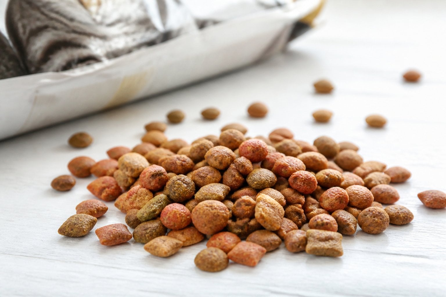 Close-up of dry cat food kibble scattered on a light surface with a blurred bag of cat food in the background.